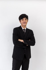 Obraz na płótnie Canvas A young asian businessman in a black suit and white shirt, standing against a white background smiling towards camera. Portrait of handsome man smiling with arms crossed on isolated white background.