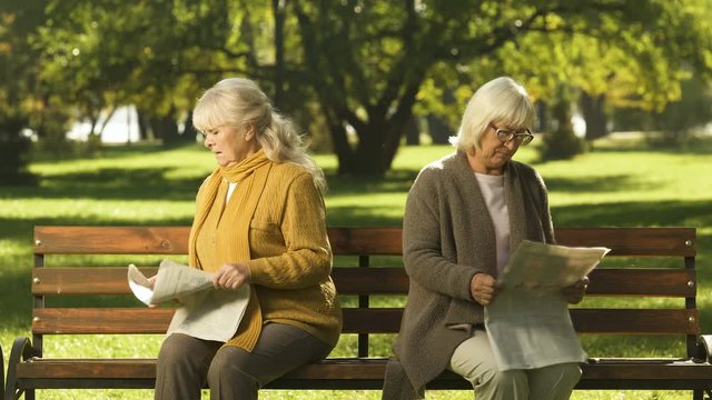 Two senior offended female friends reading newspapers, sitting on bench in park