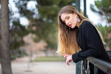 Side view of attractive young woman in elegant outfit leaning on fence and looking away while standing on blurred background of park