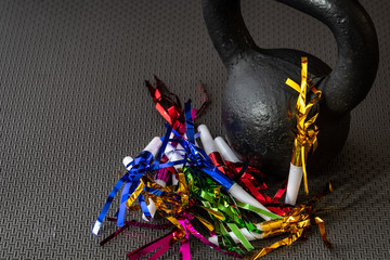 Black kettlebell on a black gym floor with blue, green, red, pink and gold noisemakers to celebrate New Year’s Eve fitness