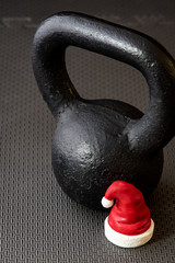 Naklejka na ściany i meble Black kettlebell on a black gym floor with small red and white Santa Claus hat to celebrate Christmas fitness