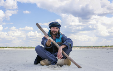 Man In The Ethnic Clothes Shows A Sand By Hand Sitting In A Desert