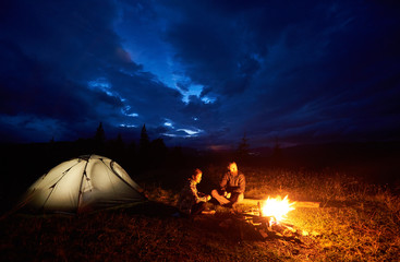 Fototapeta na wymiar Young couple man and woman tourists enjoying at night camping in the mountains, sitting near burning campfire and illuminated tourist tent under beautiful evening cloudy sky. Tourism concept