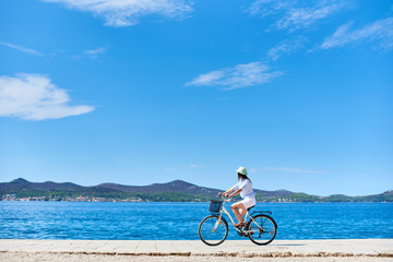 Fototapeta premium Girl cyclist in white summer closing and hat riding a bicycle along stony sidewalk on blue sparkling sea water and resort town at foot of mountains on opposite shore background. Tourism and vacations.