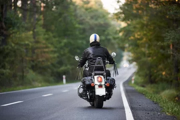 Photo sur Plexiglas Moto Vue arrière du motard en veste de cuir noir et casque blanc à moto le long d& 39 une route vallonnée entre de grands arbres verts. Mode de vie actif, amour du concept d& 39 aventures.