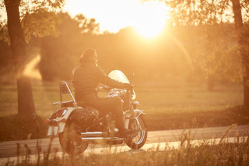 Side view of bearded motorcyclist riding modern powerful cruiser motorcycle along empty narrow country road at sunset on beautifull golden autumn landscape background.