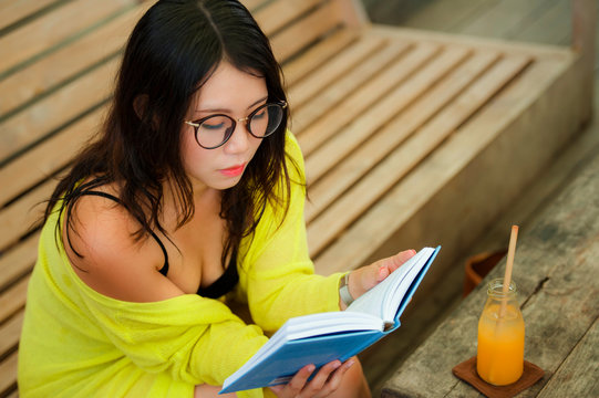 candid lifestyle portrait of young beautiful and relaxed Asian Korean student girl on reading book or studying outdoors at coffee shop drinking orange juice in nerd glasses