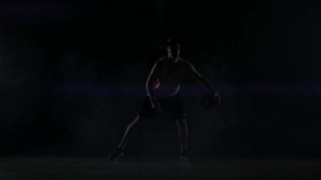 A basketball player dribbles down the court out of the shadows