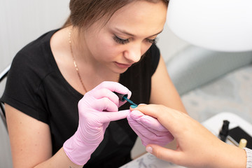 In the nail salon, a manicurist in gloves covers client's nails with a green nail Polish with a brush