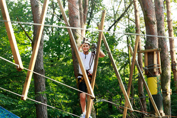 Fototapeta premium Cute boy enjoying activity in climbing adventure park at sunny summer day. Kid climbing in rope playground structure. Safe climbing with helmet insurance. Child in forest adventure park, extreme sport