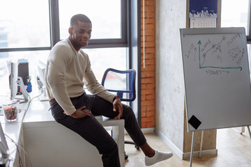 Portrait of young African American male student wearing white pullover and formal trousers, sitting inside university campus, looking at camera with pleased smile, happy to pass exams