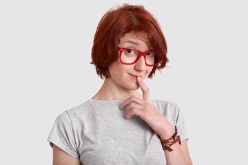 Curious red haired young woman keeps fore finger at lips, looks with interest, wears red rimed...