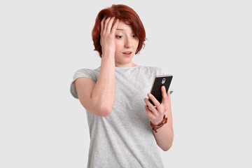 Fototapeta premium Photo of frustrated redhead woman keeps hand on head, holds modern cell phone, surfes social networks, connected to wireless internet, puzzled with latest news, isolated over white background