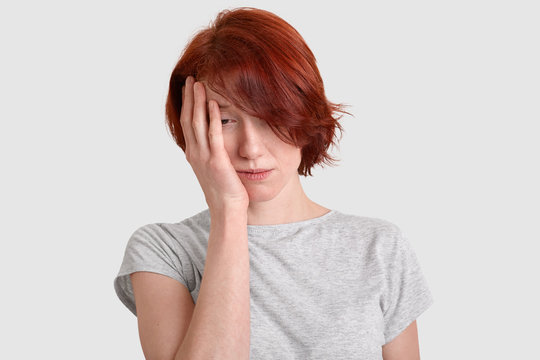 Sleepy sad woman with short red hair, feels depressed, dissatisfied, dressed in white t shirt, feels overworked after much work, isolated over white studio wall. European woman suffers from migraine