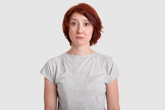 Dejected miserable woman with red hair, minimal make up, looks in displeasure at camera, wears casual grey t shirt, expresses negative emotions, feels sad and unhappy, isolated on white studio wall
