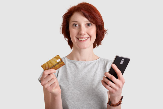Horizotnal shot of pleased woman with gentle smile, foxy hair, holds mobile phone and credit card, makes money transactions, transfers money, enjoys online shopping, checks her bank account.