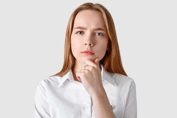 Headshot of thoughtful serious woman with blue eyes, soft skin, holds chin and looks directly at camera, has long straight hair, dressed in elegant shirt, isolated over white background. Thoughts