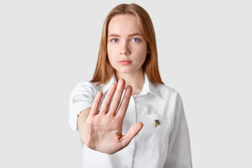 Young lady makes stop gesture with palm, has serious facial expression, dressed in elegant white shirt, refuses to speak with boyfriend after quarrel, models over white studio background. Rejection