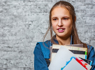 portrait of young teenager brunette girl with long hair holding books and note books wearing backpack on gray wall background