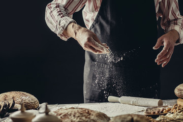 Male Cook hands kneading dough, sprinkling piece of dough with white wheat flour. Low key shot, close up on hands, tasty rustic organic loaf of bread and some ingredients around on table.