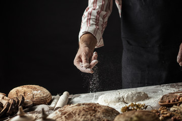 The process of making home bread by male hands. Powdery flour flying into air as man in black chef outfit sprinkles it on dough over white table covered in flour