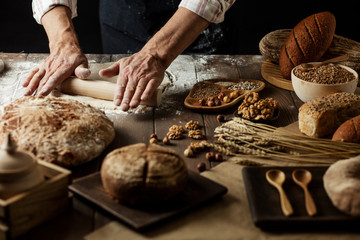 Hands kneading raw dough on table. Top view on baker workplace, working with pastry, all surface on flowered table is occupied with loaves of bread and ingredients. Culinary, cooking, bakery concept