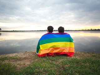 Two young man covered by a rainbow flag beside the lake on sunset sky background. LGBT, Gay, Pride...