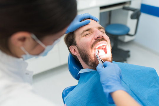 Female dentist pulling teeth out on a male patient with open mouth sitting in dentist chair in a modern dental clinic. Pain, tooth extraction, dentistry concept.