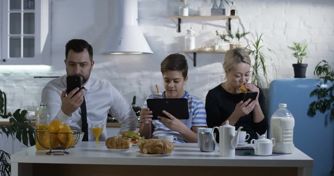 Medium shot of a family sitting side by side at the kitchen table and watching their smartphones and tablets