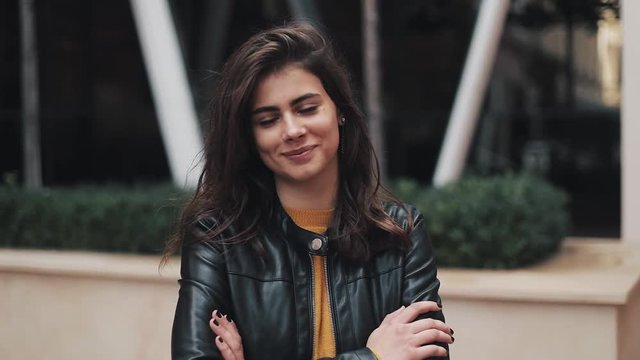Portrait shot of the young attractive woman standing near office building smiling to the camera with hands crossed in front of her. She is dressed in a warm yellow sweater and autumn leather jacket