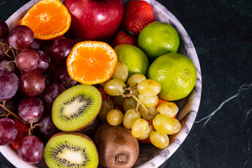 Fresh tropical fruitson in bowl on black marble background. Pineapple, papaya, mango, kiwi, grape, garnet, tangerines, bananas, apples, watermelon slices, pitaya or dragon fruit