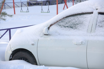 The front part of the white car is covered with a layer of snow. Parked car in the snow-covered Parking lot.