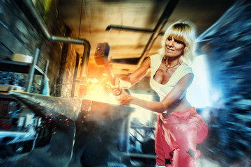 Fototapeta na wymiar Woman blacksmith forges metal on the anvil with the sledgehammer. Workshop on the background.