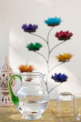 Glass pitcher with water and drinking glasses surrounded by chakra flowers and peaceful elements.