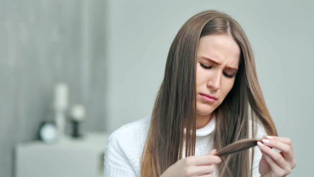 Upset young Caucasian woman looking on weak unhealthy dry hair ends at home interior