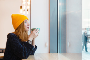 Young woman with blue eyes and blond hair in a yellow knitting hat is drinking coffee in a cafe and looking into the window.