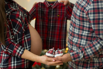 Future parents holding hands and a pair of little shoes and a boy shirt over christmas background. Concept of Parents-To-Be.
