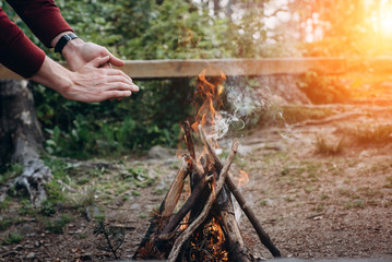 Fototapeta premium Summer camp. Hands man near a campfire at sunset time in the forest . Concept adventure active vacations outdoor.