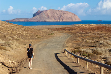 Young female hiking on a dirt road towards the sea in a volcanic landscape on a hot summer day....