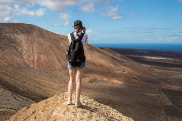 Young Caucasian woman with a backpack, wearing shorts, hiking on the Caldera Blanca volcano in Lanzarote and taking pictures amazed by the surrounding landscape.