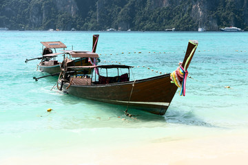 Long boats stand on the island of Phi Phi. Clear blue water can be seen.