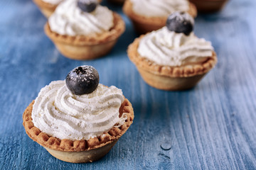 Bright tartlets filled with curd cream and decorated with blueberries on top are on a blue wooden desk, close up