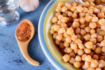 Boiled chickpeas, seasoned with vegetable oil, ground red pepper, sesame seeds and onion rings in a bowl with old spoon on a blue old wooden desk next to red hot pepper and garlic, top view