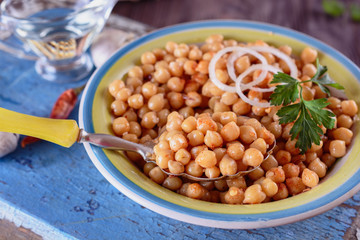 Boiled chickpeas, seasoned with vegetable oil, ground red pepper, sesame seeds and onion rings in a bowl with old spoon on a blue old wooden desk next to red hot pepper and oil, close up