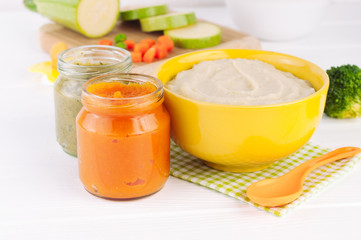 Different types of vegetable puree in bowl and jars on white wooden background