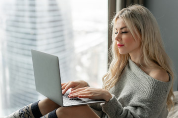 Girl typing on the keyboard. Beautiful blonde woman sitting on the bed and looks at the laptop screen. Blogging, browsing internet, chatting. In a warm cozy sweater and wool socks.