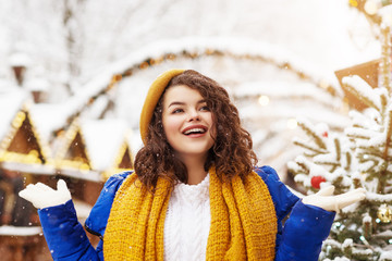 Outdoor portrait of young happy smiling woman catching snow, posing at festive Christmas fair in...