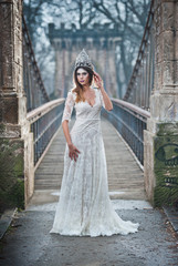 Lovely young lady wearing elegant white dress enjoying the beams of celestial light and snowflakes falling on her face. Pretty brunette girl in long wedding dress posing on a bridge in winter scenery