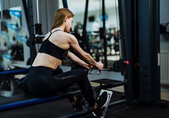 Fototapeta na wymiar Muscular young woman in black sportwear and trainers is doing intensive workout by pulling weights at the gym. Athletic female is doing muscle training on a simulator in the sport club.