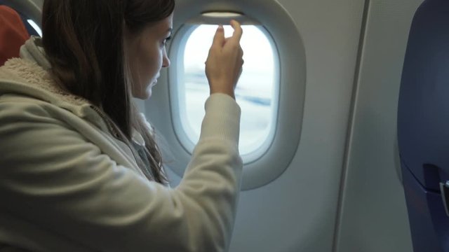 Beautiful woman is opening porthole and looking out from window in airplane